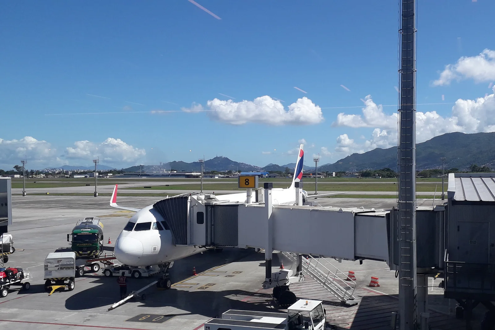 Avião parado numa ponte de embarque do Aeroporto Internacional de Florianópolis, com o pátio e os morros ao fundo.