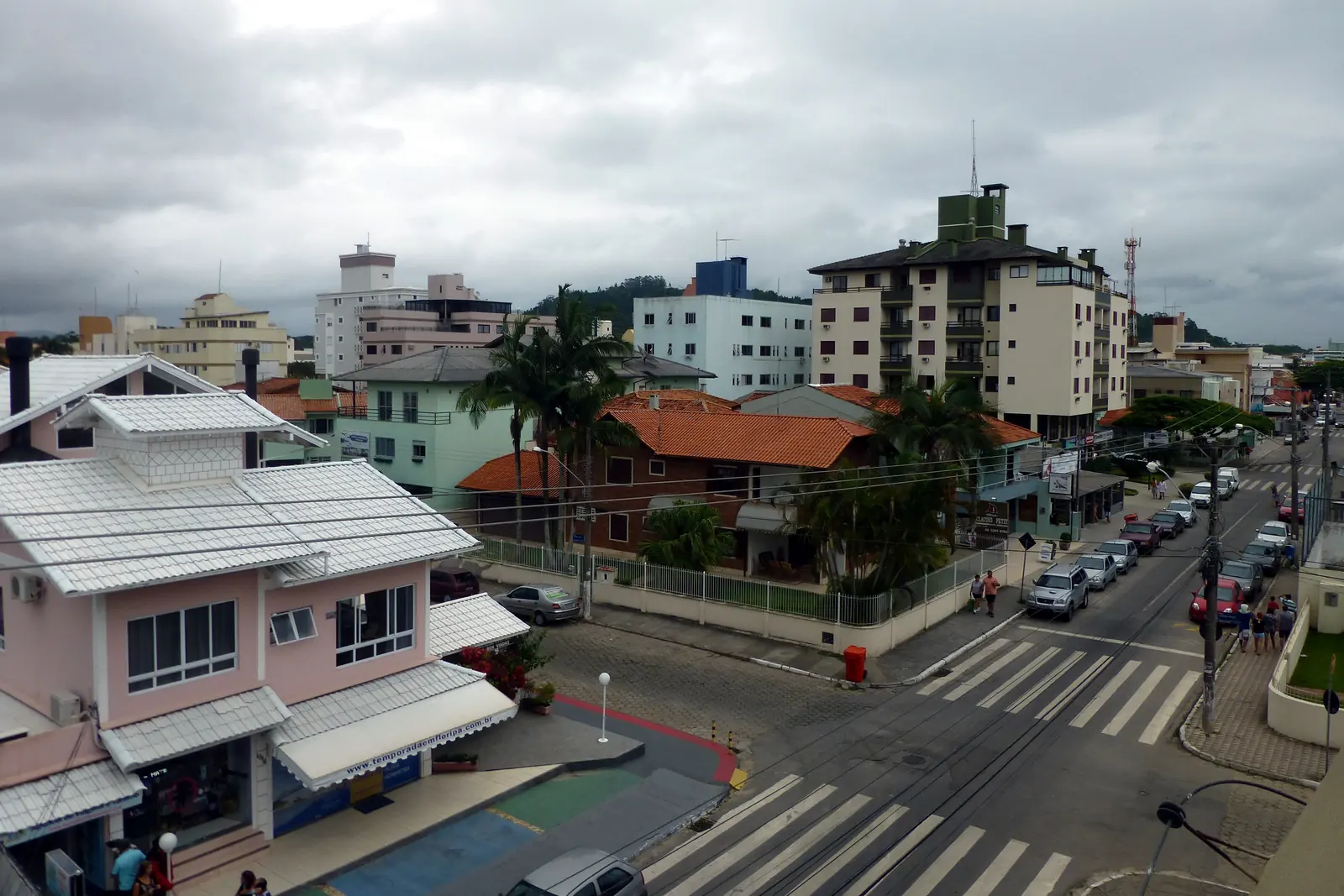 Rua Maria Villac, em Canasvieiras, no norte da ilha, vista do alto num dia nublado: casas e prédios baixos, carros estacionados e faixas de pedestres.